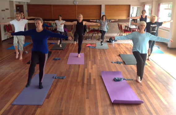 A class balancing on one leg with arms out, in a mirrored hall with a wooden floor